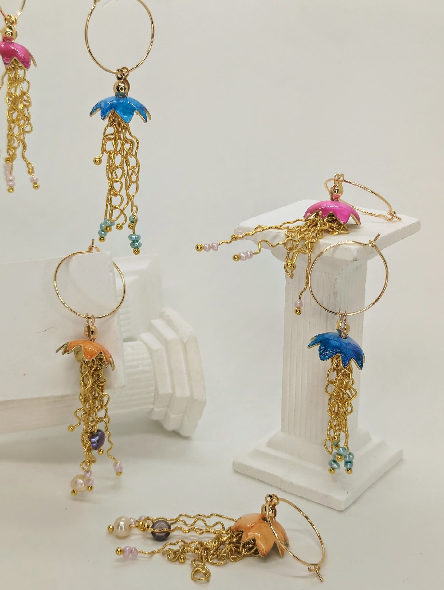 A collection of jelly fish inspired earrings, displayed on white columns, some jelly fish earrings are hanging in mid air. The jelly fish are various colours including blue, orange and pink with a cascade of gold plated tentacles with freshwater pearls