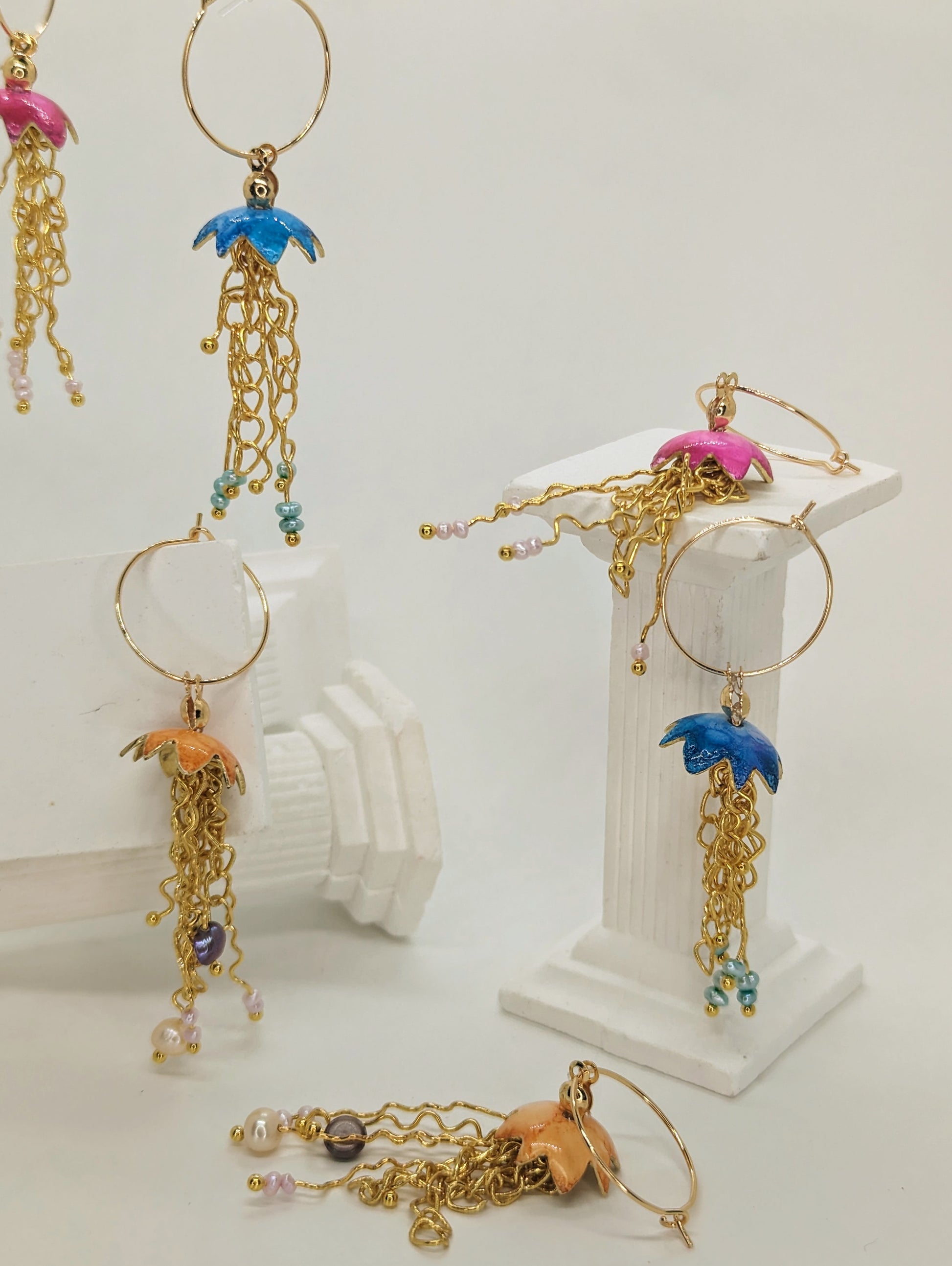 A collection of jelly fish inspired earrings, displayed on white columns, some jelly fish earrings are hanging in mid air. The jelly fish are various colours including blue, orange and pink with a cascade of gold plated tentacles with freshwater pearls