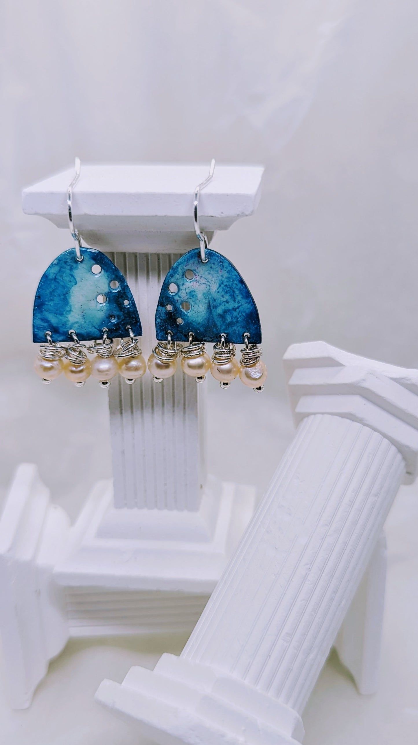 Ocean blue earrings, enamelled aluminium with different shades of blue embellished with four apricot freshwater pearls on each earring. The earrings are hanging from sterling silver hooks. All displayed on white columns.
