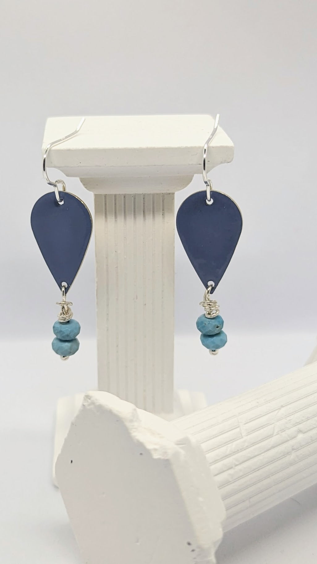 Earrings displayed on white columns. Enamelled tear drop brass in a blue colour embellished with two turquoise stones each. The earrings are hanging from sterling silver earring hooks.