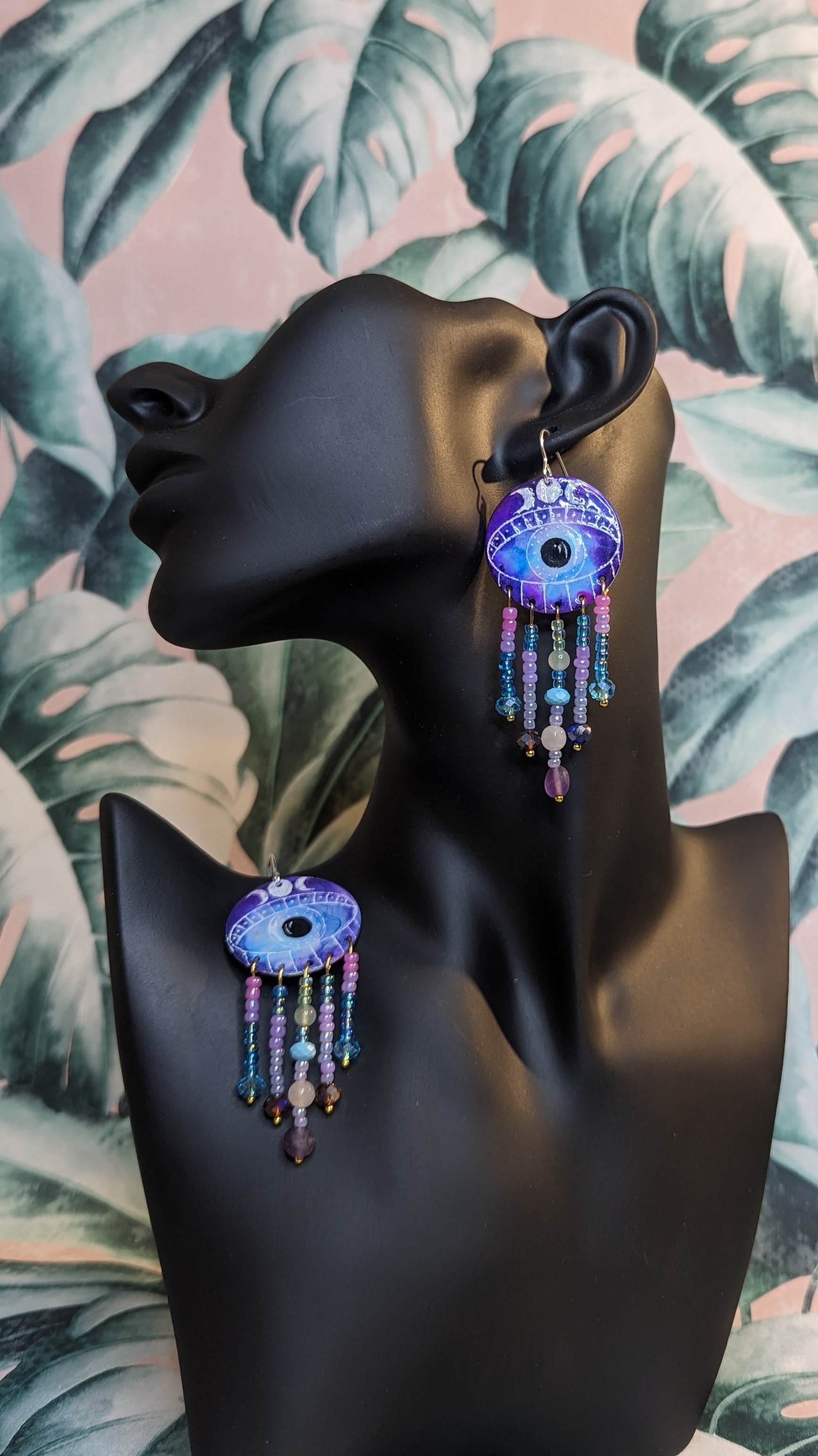 Earrings. Medium Circular aluminium disc enamelled with a mystical colour combination with a large eye in the centre with a crescent moon shapes above the eyelid. Five gold plated pins hang from the eye, embellished with acrylic beads, glass crystals and healing crystals. including Amethyst, green aventurine, rose quartz and turquoise. The earrings are displayed on a black mannequin with a floral background.