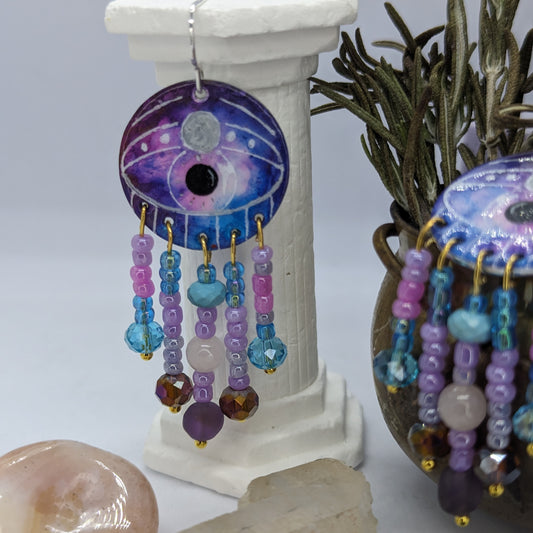 Earrings. Circular aluminium disc enamelled with a mystical colour combination with a large eye in the centre with a moon shape on the eyelid. Five gold plated pins hang from the eye, embellished with acrylic beads, glass crystals and healing crystals. including Amethyst, rose quartz and turquoise.