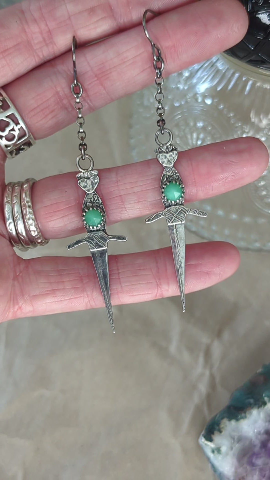 Athame witch's ritual dagger earrings hanging from a jeweller's hand, gently swaying with a crystal tea cup in the background. The details of the oxidised silver and chrysoprase stones are clearly visible.