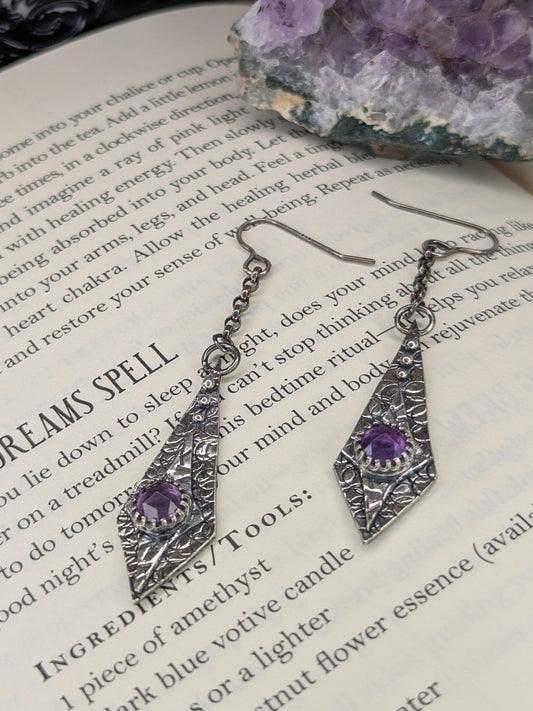 Maiden Earrings placed on an open spell book, with a raw amethyst stone in the background, creating a mystical and enchanting atmosphere.