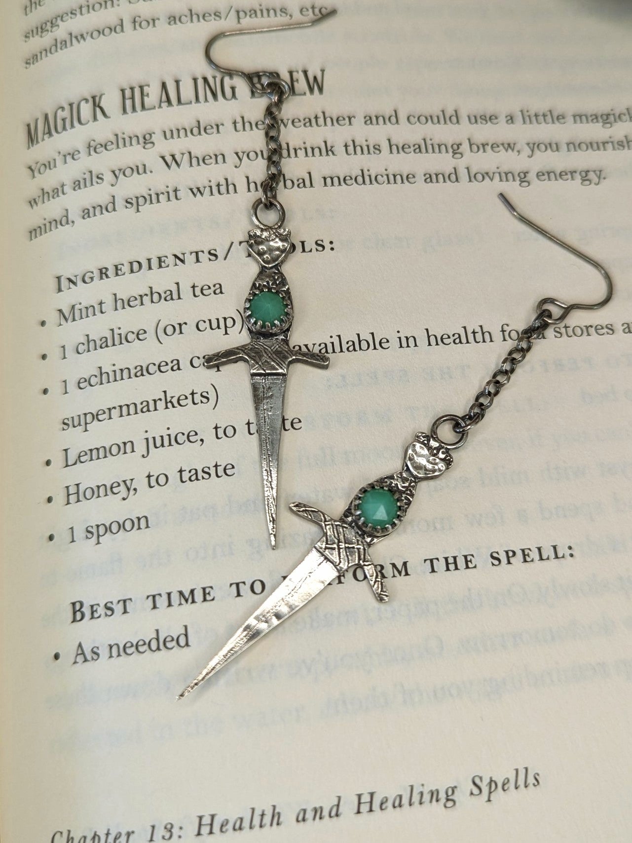 Athame witch's ritual dagger earrings displayed on the pages of an open spell book, showcasing their intricate design in recycled silver with chrysoprase stones and oxidised finish.