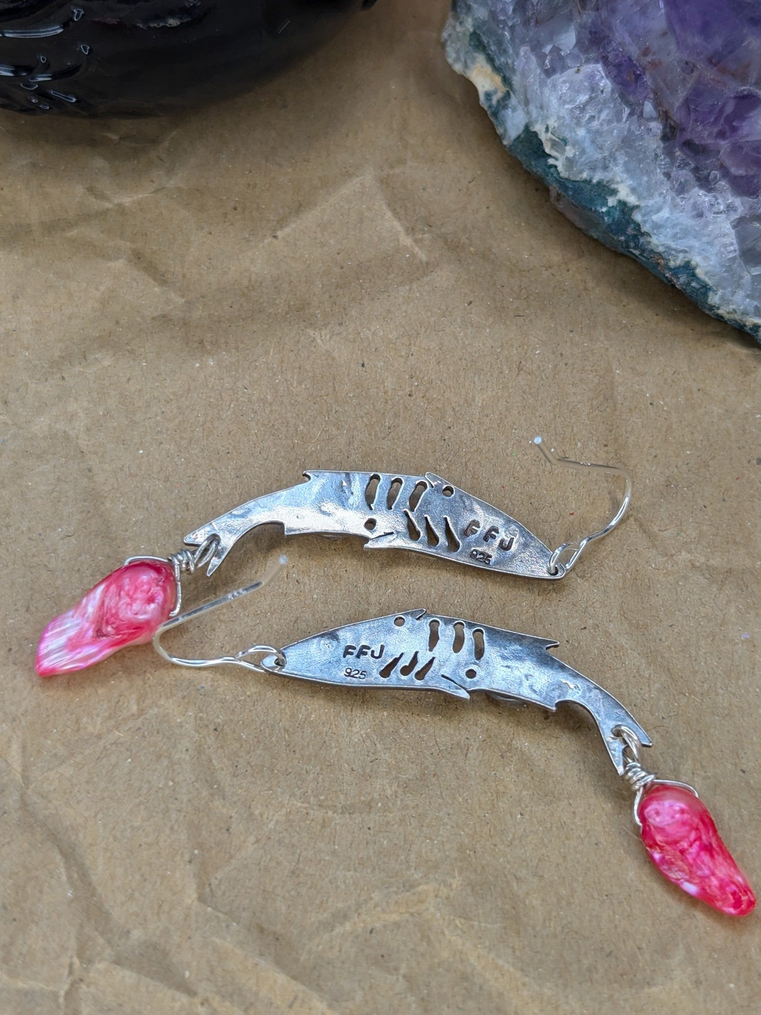 The back of the Sterling silver fish earrings, showing a 925 stamp and FFJ stamp. Pink keshi Freshwater Pearls are visible. The earrings are placed on a brown background with an amethyst stone and spell jar in the background