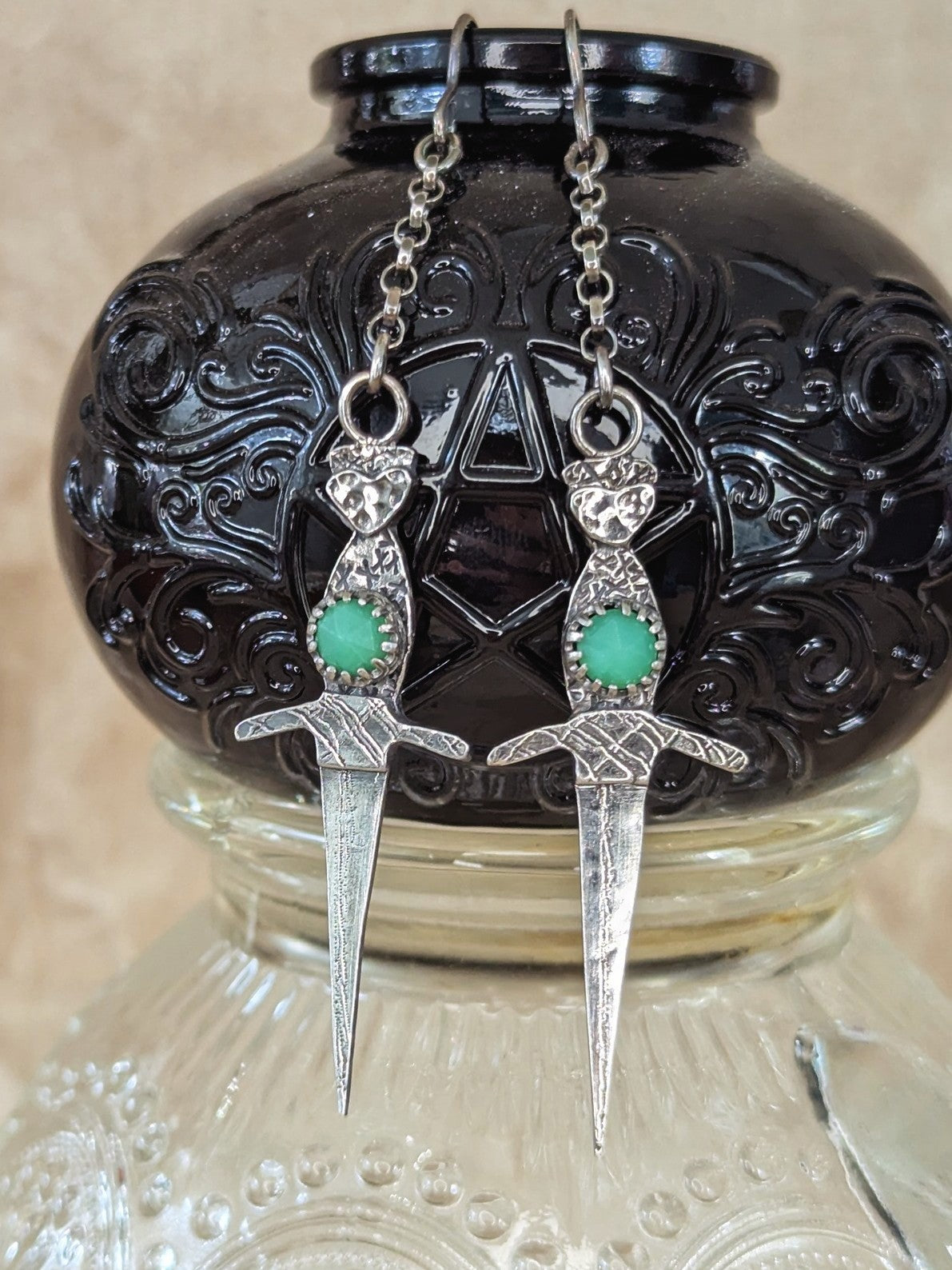Athame witch's ritual dagger earrings hanging from the edge of a purple embossed glass spell jar, highlighting the rustic, textured silver and vibrant chrysoprase stones.