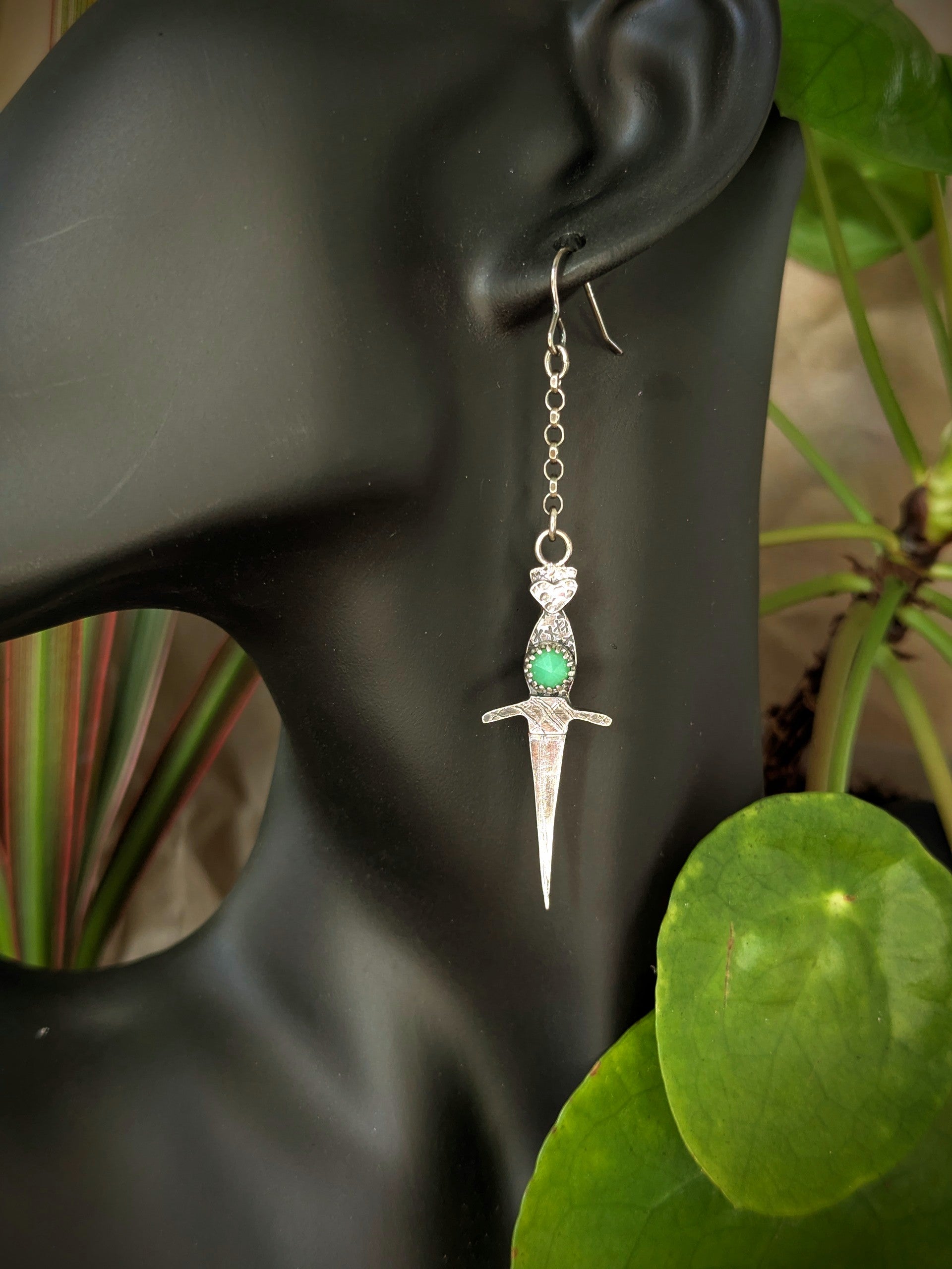 Athame witch's ritual dagger earrings displayed on a black mannequin's ear, with lush green plants softly blurred in the background, showcasing the jewellery’s vintage charm against a natural backdrop.