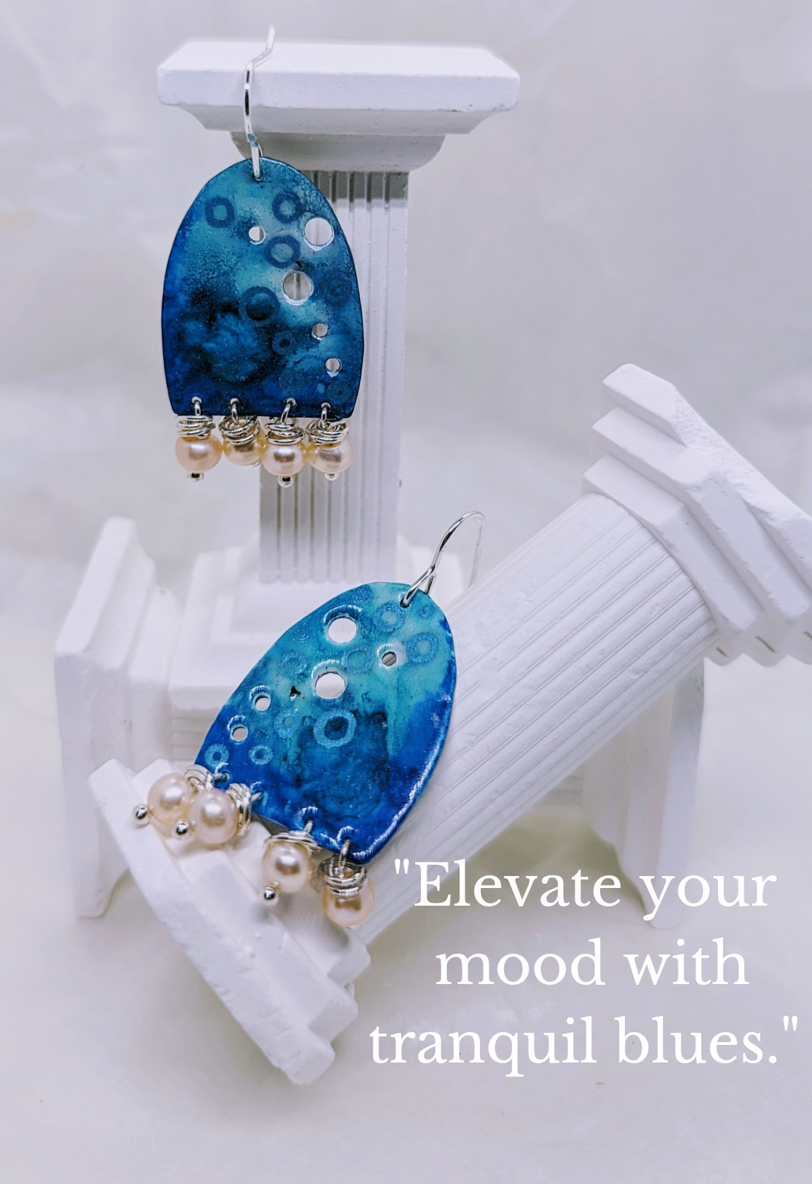 Calming blue, with an ocean wave design, embellished with apricot freshwater pearls. Enamelled aluminium for a lightweight design. The earrings are displayed on white columns. The writing on the image says "Elevate your mood with tranquil blues"  