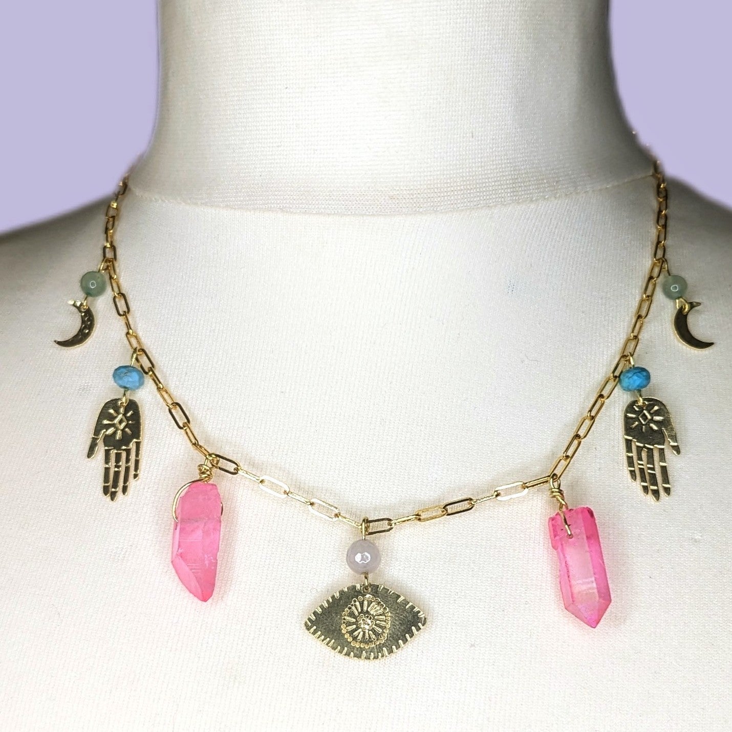 Self love potion necklace, displayed on white mannequin. Gold plated trace chain necklace with raw brass hand, eye and crescent moon embellishments, also featuring healing crystals beads including pink quartz, turquoise and green aventurine 