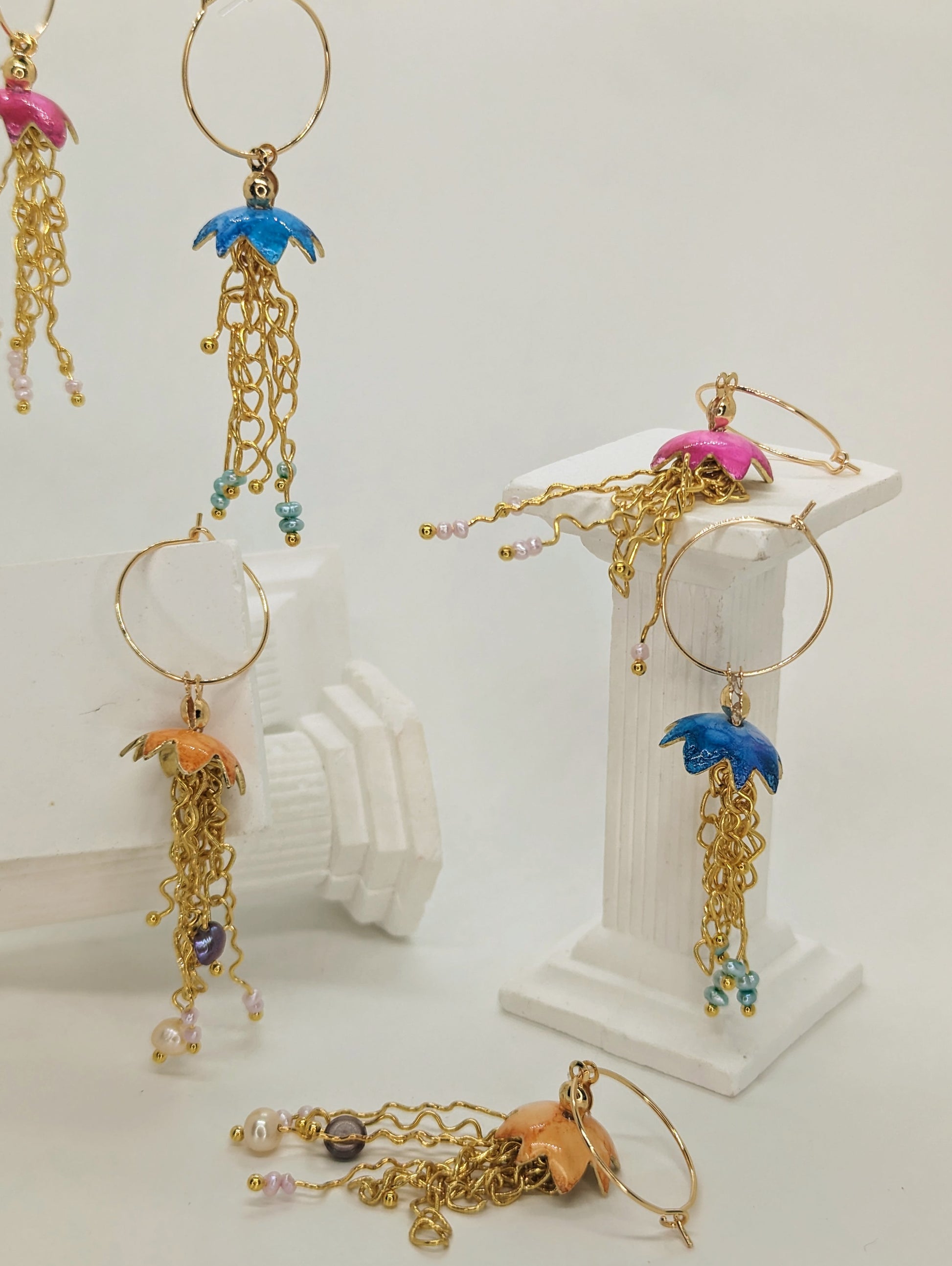 A collection of jelly fish inspired earrings, displayed on white columns, some jelly fish earrings are hanging in mid air. The jelly fish are various colours including blue, orange and pink with a cascade of gold plated tentacles with freshwater pearls 