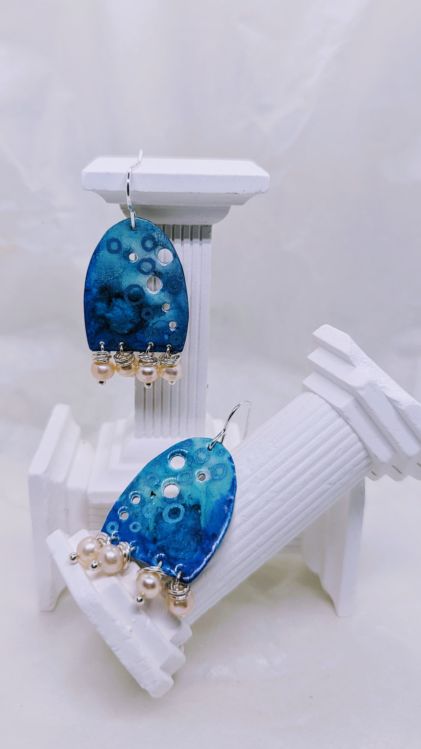 Large Ocean blue earrings, enamelled aluminium with different shades of blue embellished with four apricot freshwater pearls on each earring. The earrings are hanging from sterling silver hooks. All displayed on white columns. 