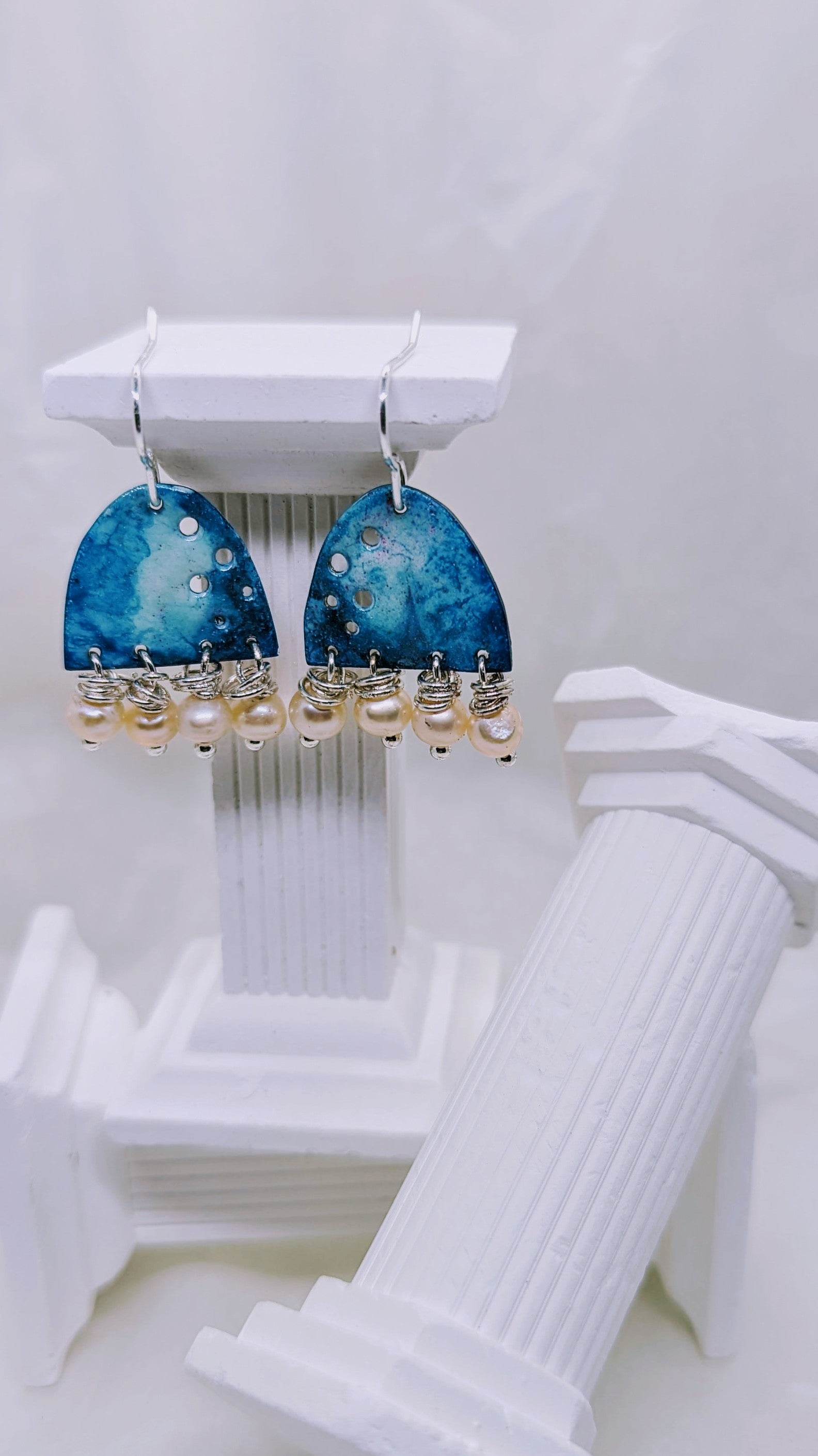 Ocean blue earrings, enamelled aluminium with different shades of blue embellished with four apricot freshwater pearls on each earring. The earrings are hanging from sterling silver hooks. All displayed on white columns. 