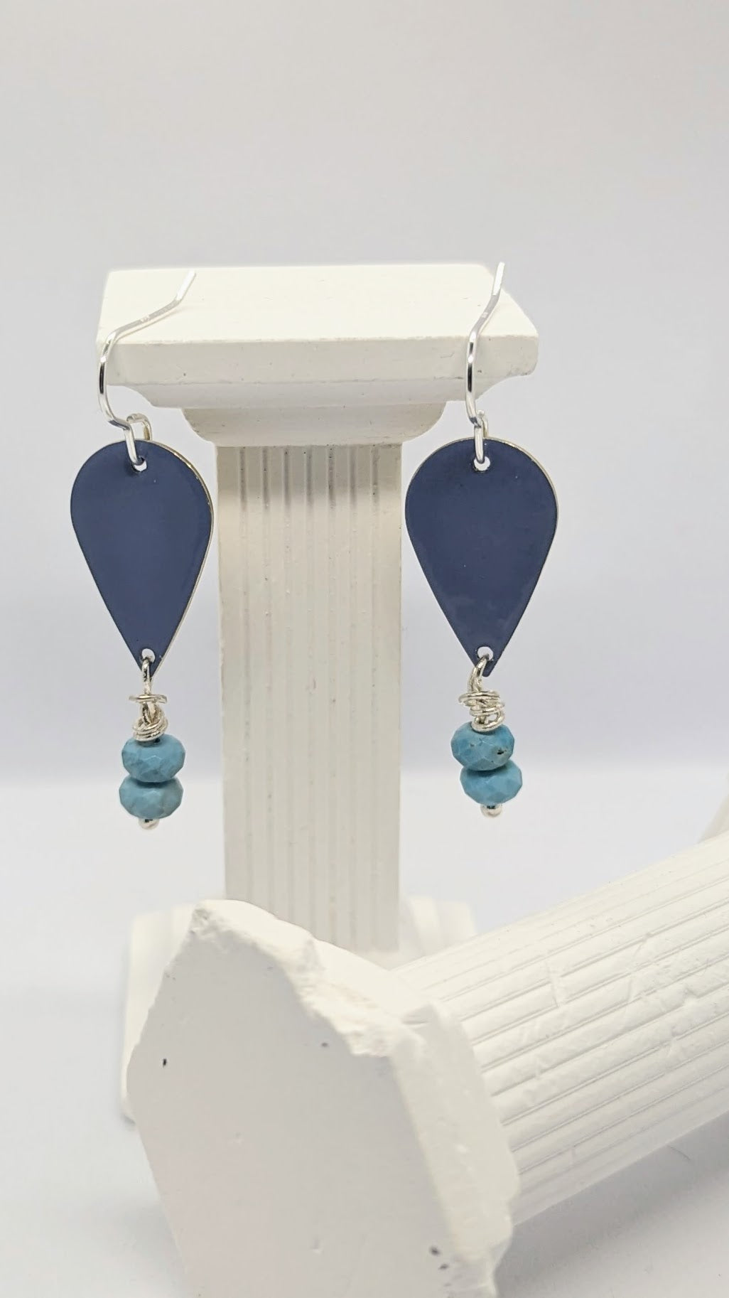 Earrings displayed on white columns. Enamelled tear drop brass in a blue colour embellished with two turquoise stones each. The earrings are hanging from sterling silver earring hooks.  