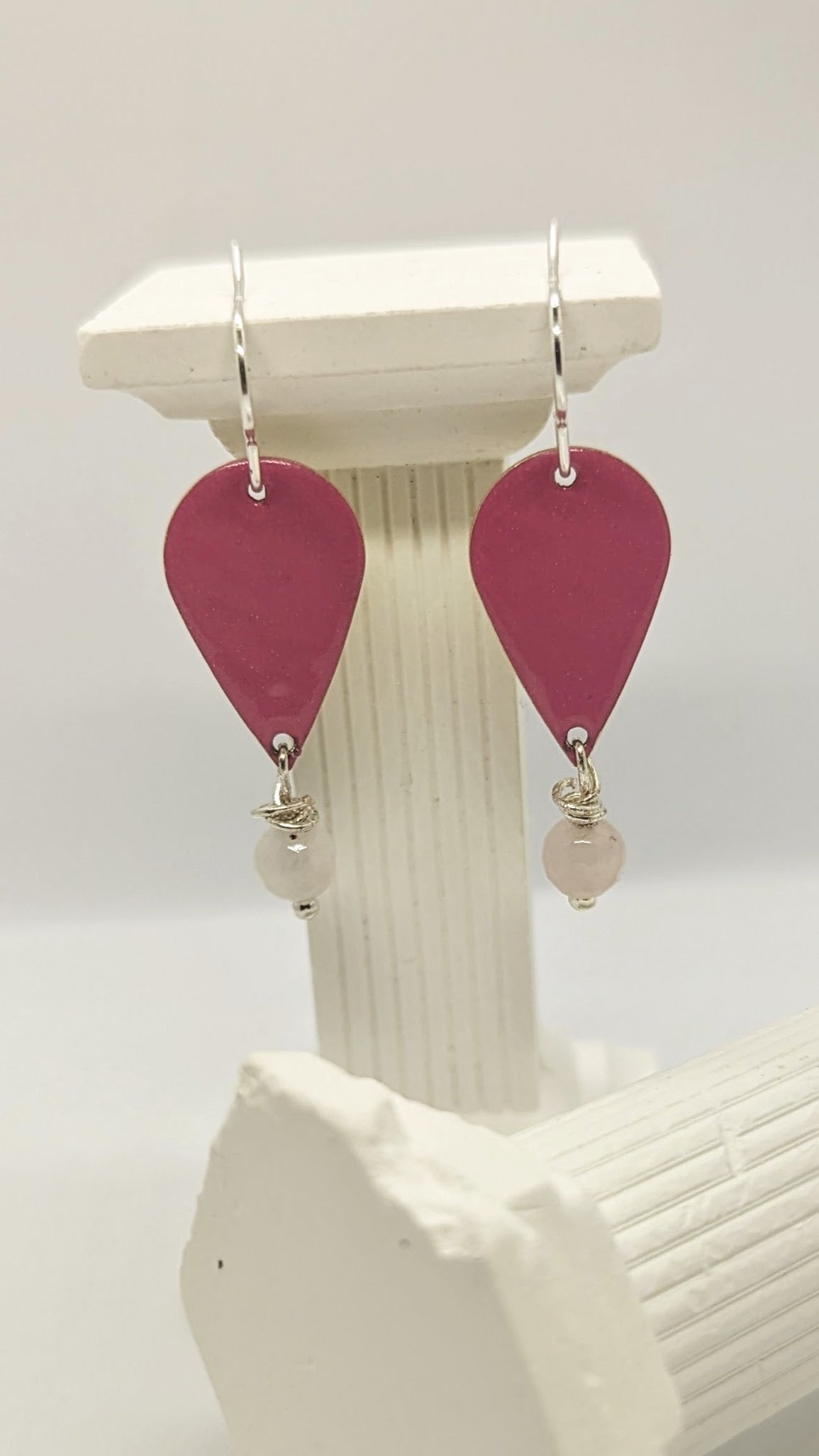 Earrings displayed on white columns. Enamelled tear drop brass in a pink colour embellished with a rose quartz each. The earrings are hanging from sterling silver earring hooks.