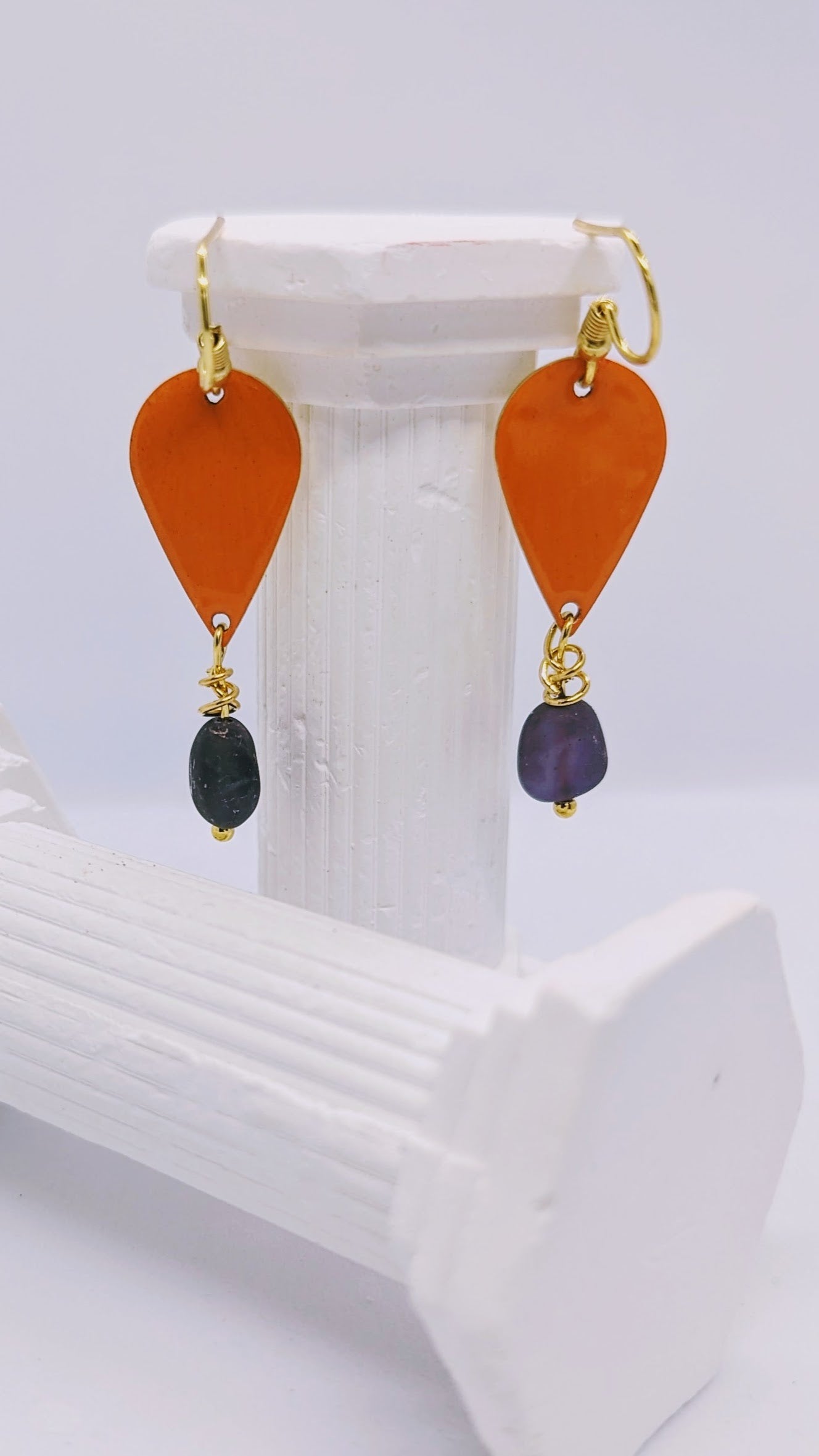 Earrings displayed on white columns. Enamelled tear drop brass in a orange colour embellished with a amethyst each. The earrings are hanging from sterling silver earring hooks.