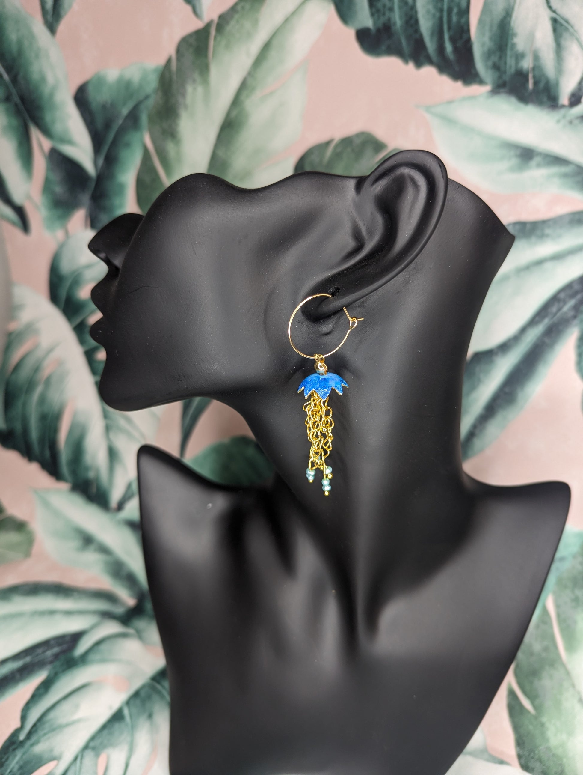 Blue Jelly fish inspired earrings with a cascade of gold plated tentacles with freshwater pearls. Displayed on a black mannequin head with a floral background 