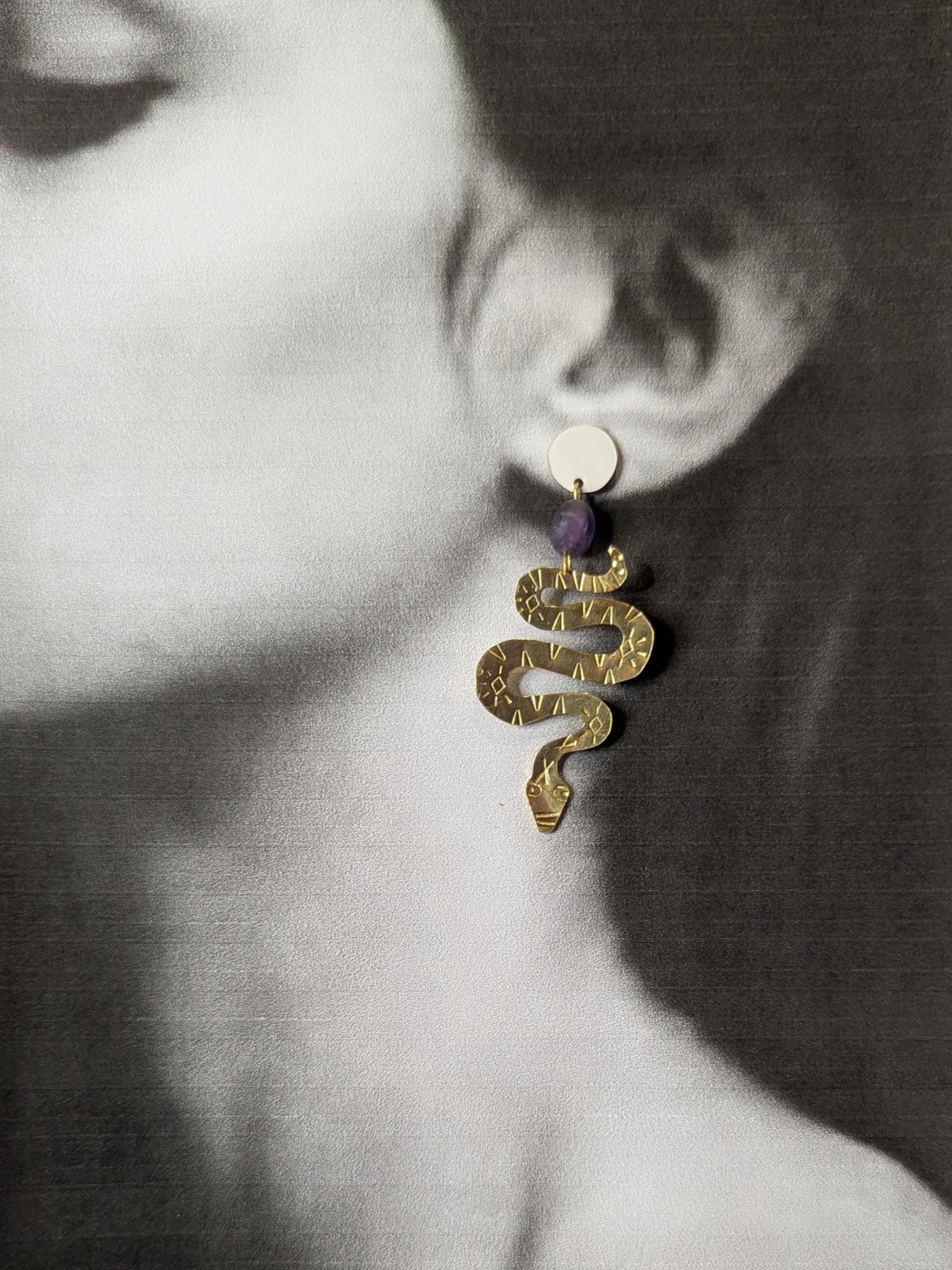 Brass textured snake earrings with amethyst beads hanging from white enamel brass studs. The earrings are placed on a black and white image of a model.