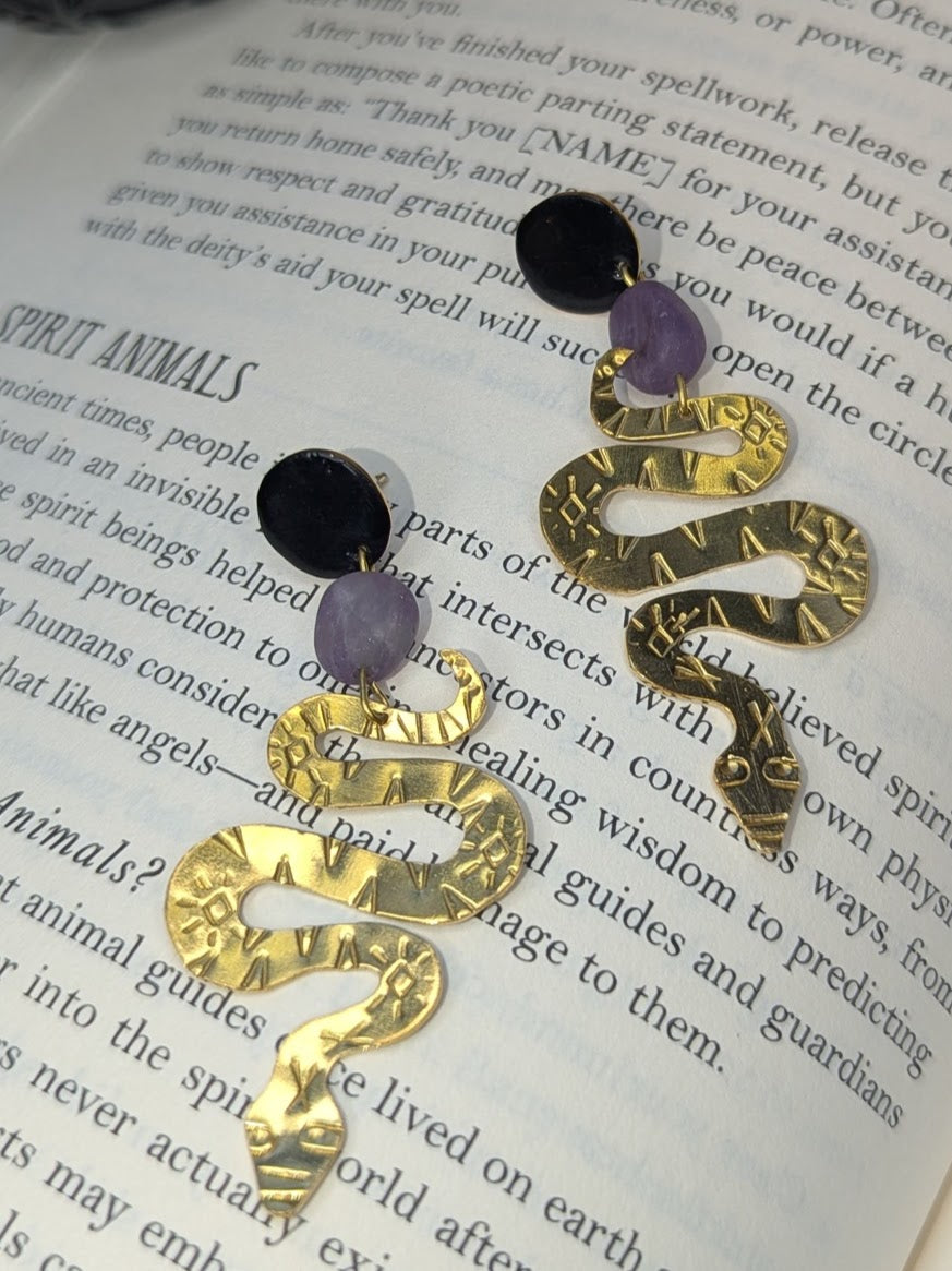 Brass textured snake earrings with amethyst beads hanging from black enamel brass studs. The earrings are placed on a spell book page talking about spirit animals.