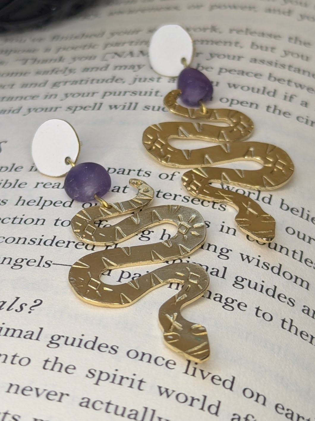 Brass textured snake earrings with amethyst beads hanging from white enamel brass studs. The earrings are placed on a spell book page talking about spirit animals.