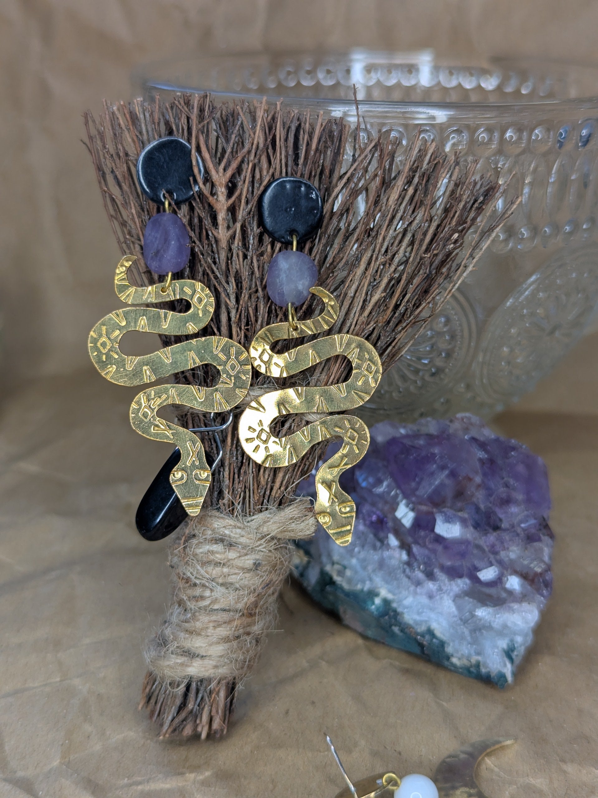 Brass textured snake earrings with amethyst beads hanging from black enamel brass studs. The earrings are hanging on altar besom that is leaning on a crystal tea cup with an amethyst crystal behind the besom.