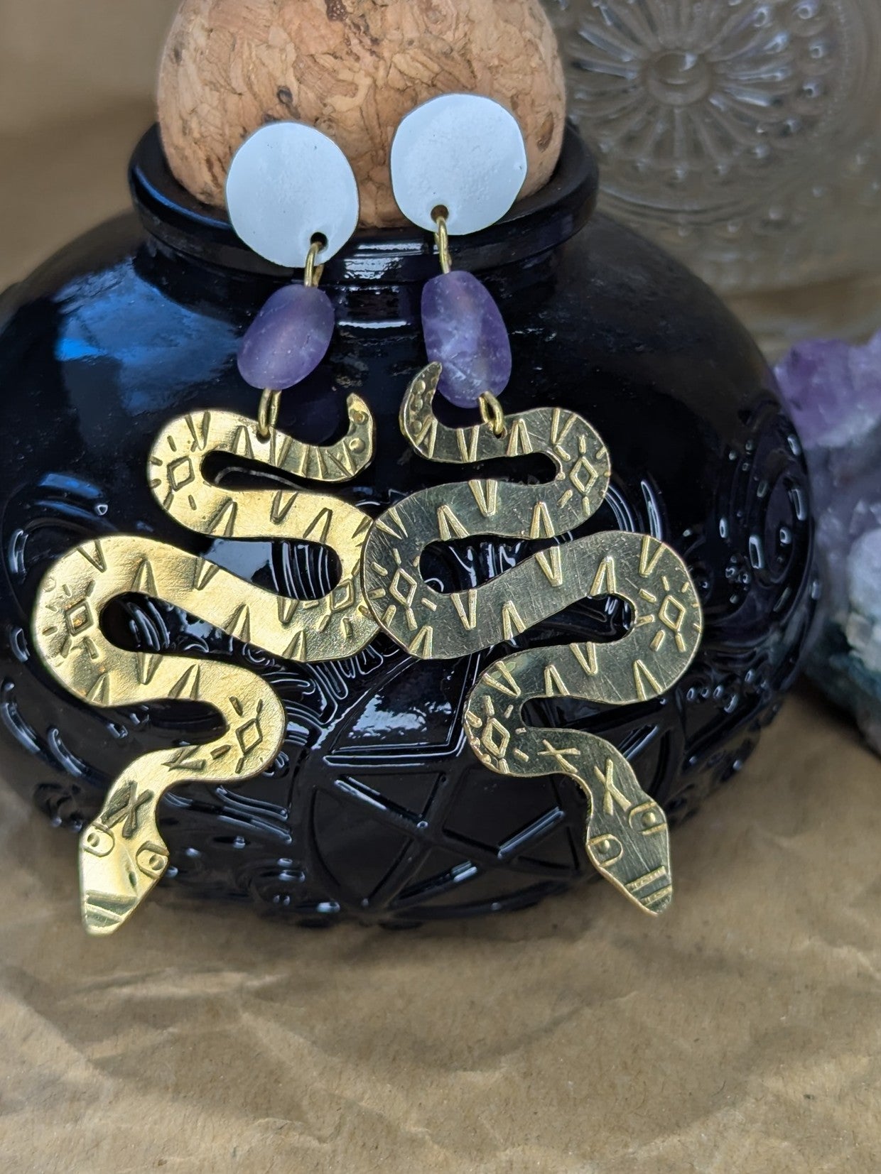 Brass textured snake earrings with amethyst beads hanging from white enamel brass studs. The earrings are hanging on a black spell bottle on top of brown packing paper, with a amethyst crystal in the background.