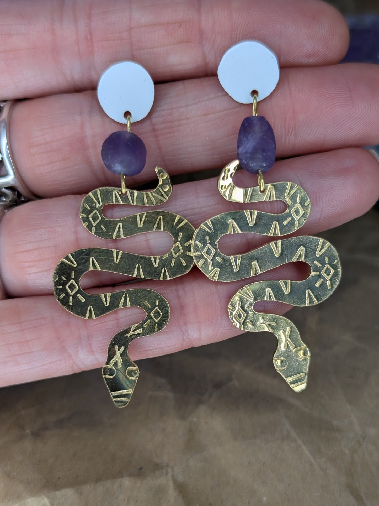 Brass textured snake earrings with amethyst beads hanging from white enamel brass studs. The earrings are placed on a hand to show the contrast with skin.