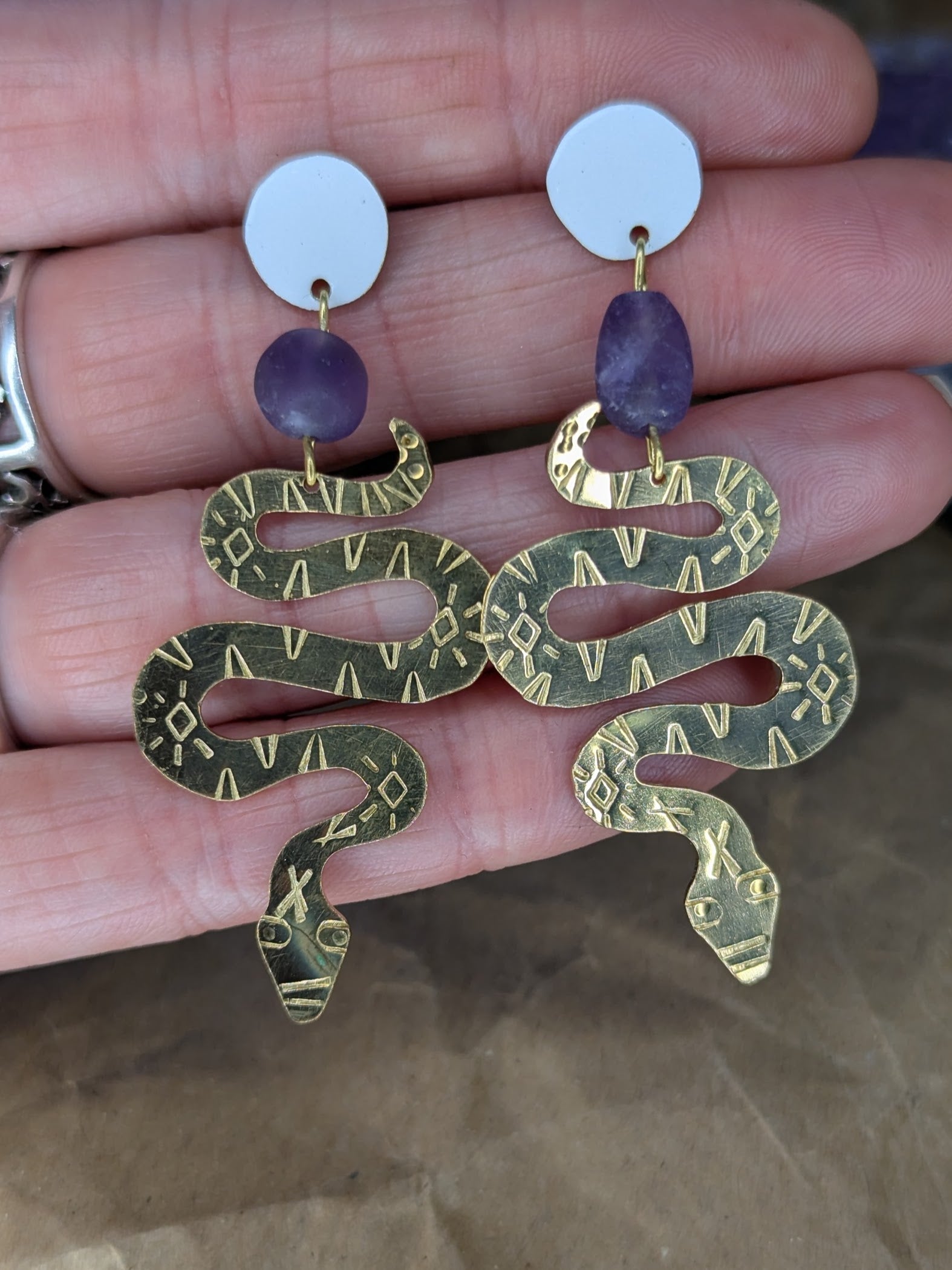 Brass textured snake earrings with amethyst beads hanging from white enamel brass studs. The earrings are placed on a hand to show the contrast with skin.