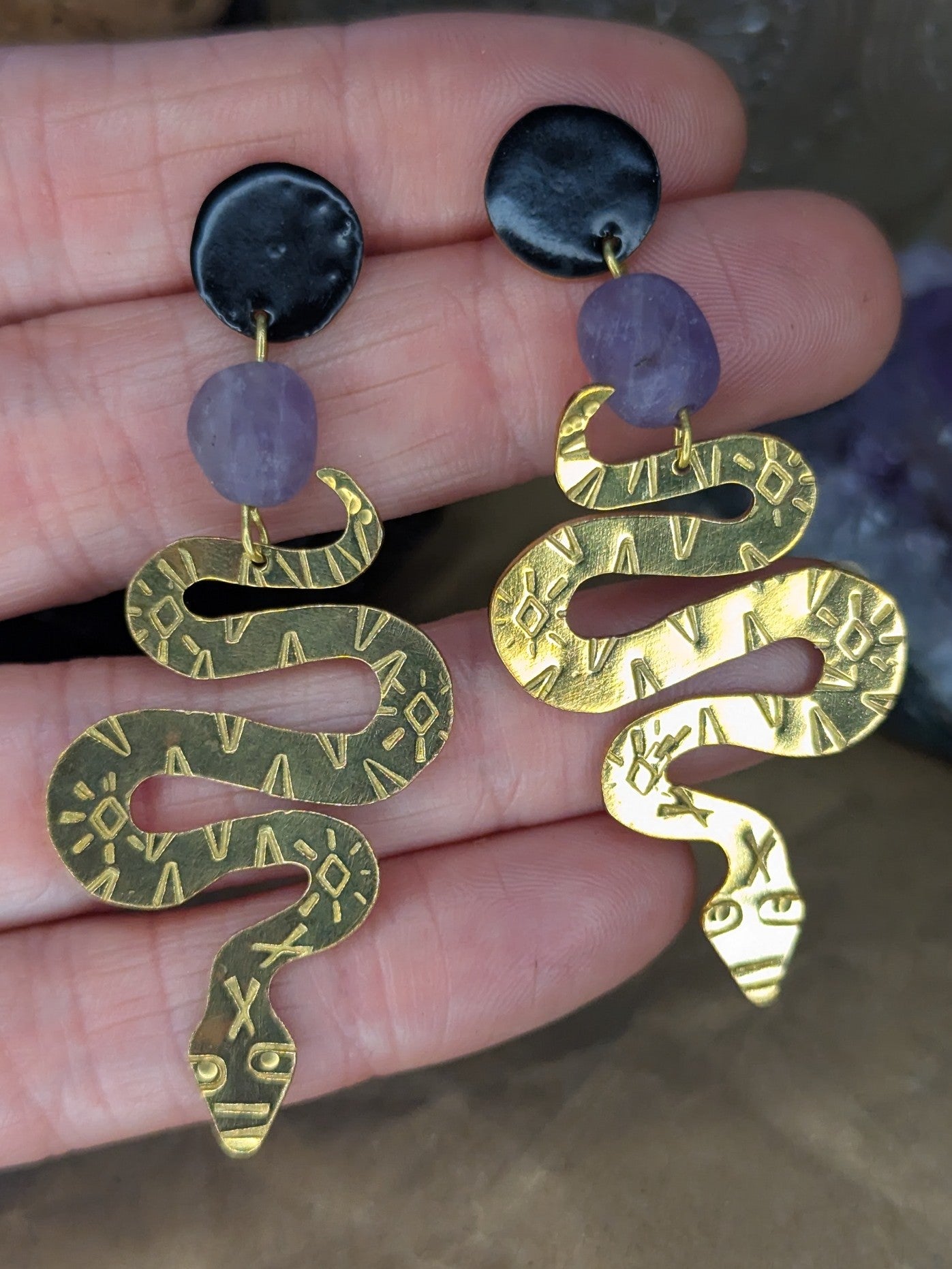 Brass textured snake earrings with amethyst beads hanging from black enamel brass studs. The earrings are placed on a hand to show the contrast with skin with a floral background.