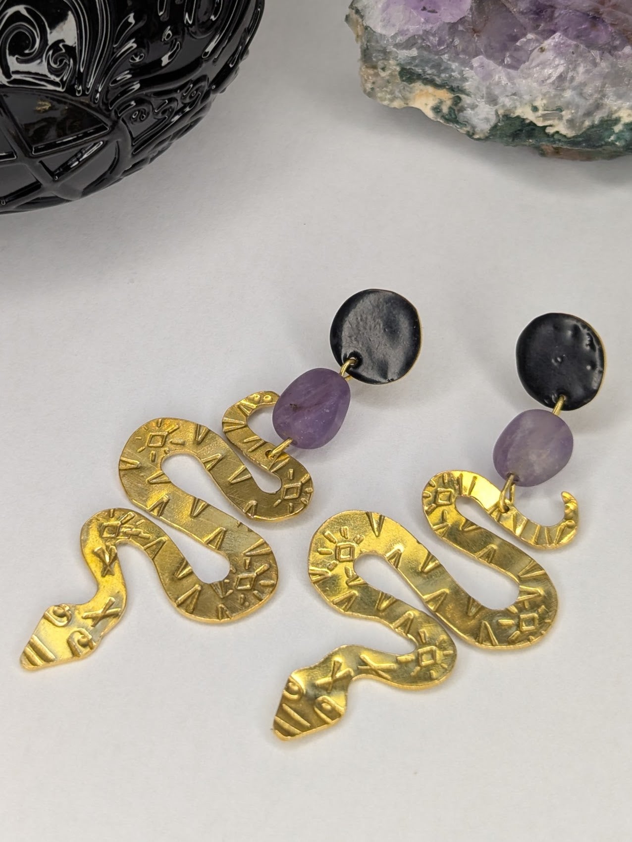 Brass textured snake earrings with amethyst beads hanging from black enamel brass studs. The earrings are placed on a white background with an amethyst crystal and spell jar in the background.