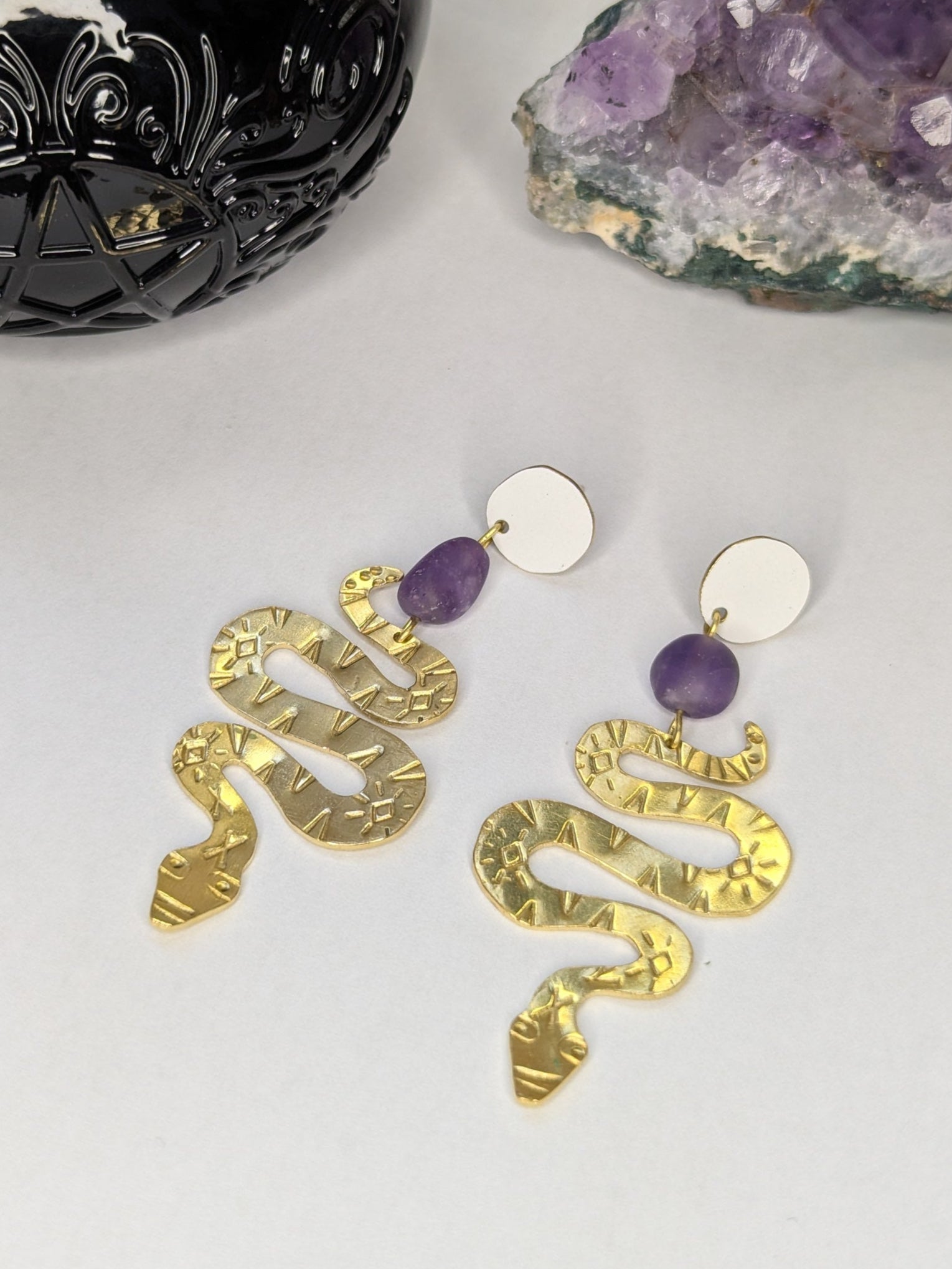 Brass textured snake earrings with amethyst beads hanging from white enamel brass studs. The earrings are placed on a white background with an amethyst crystal and spell jar in the background.